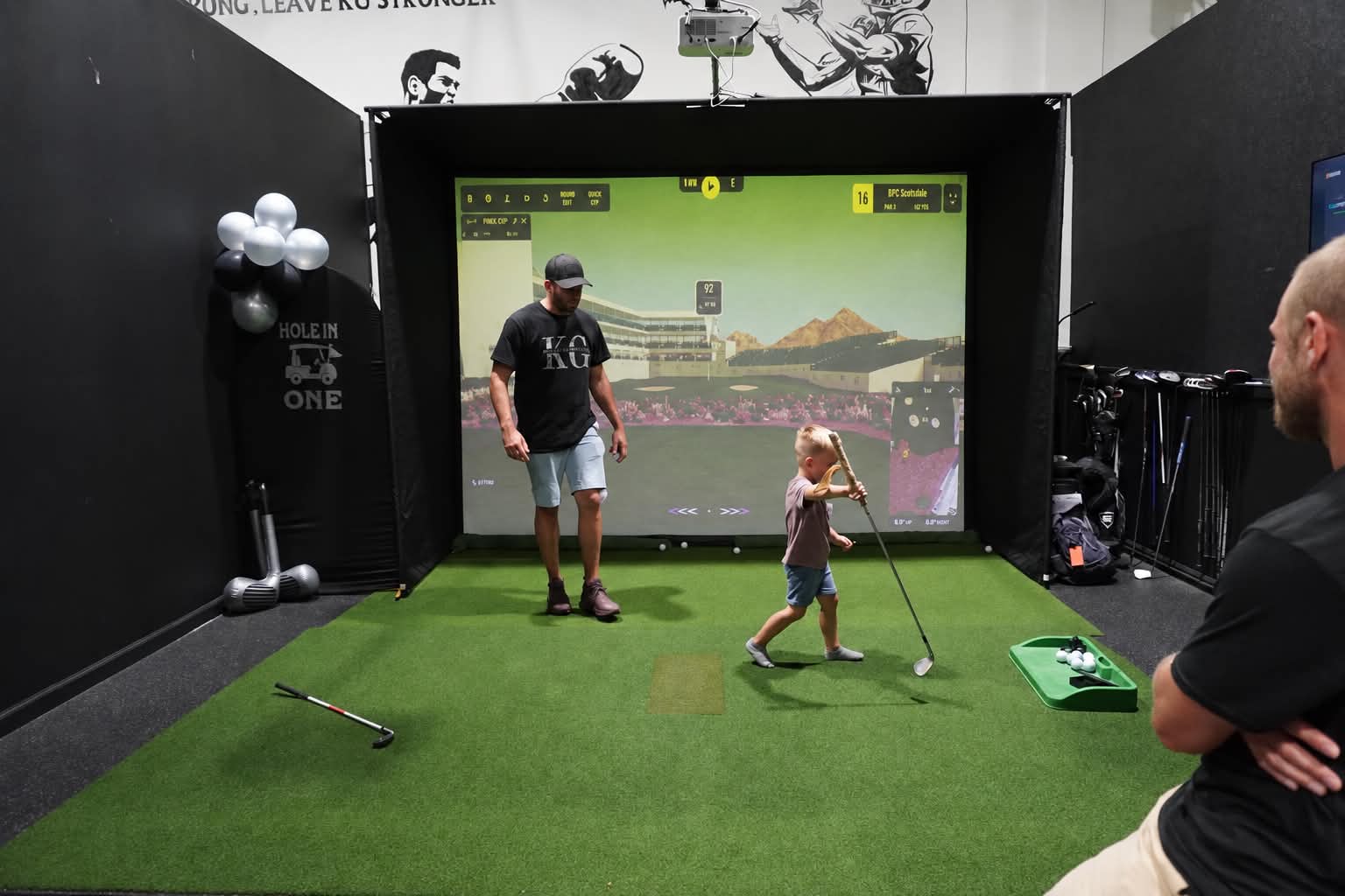 A child learning to swing in the golf simulator with family nearby